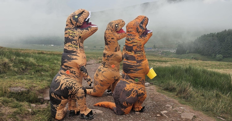 Three people in dinosaur costumes look up into the sky, the fog from the mountains in the distance. It is majestic.
