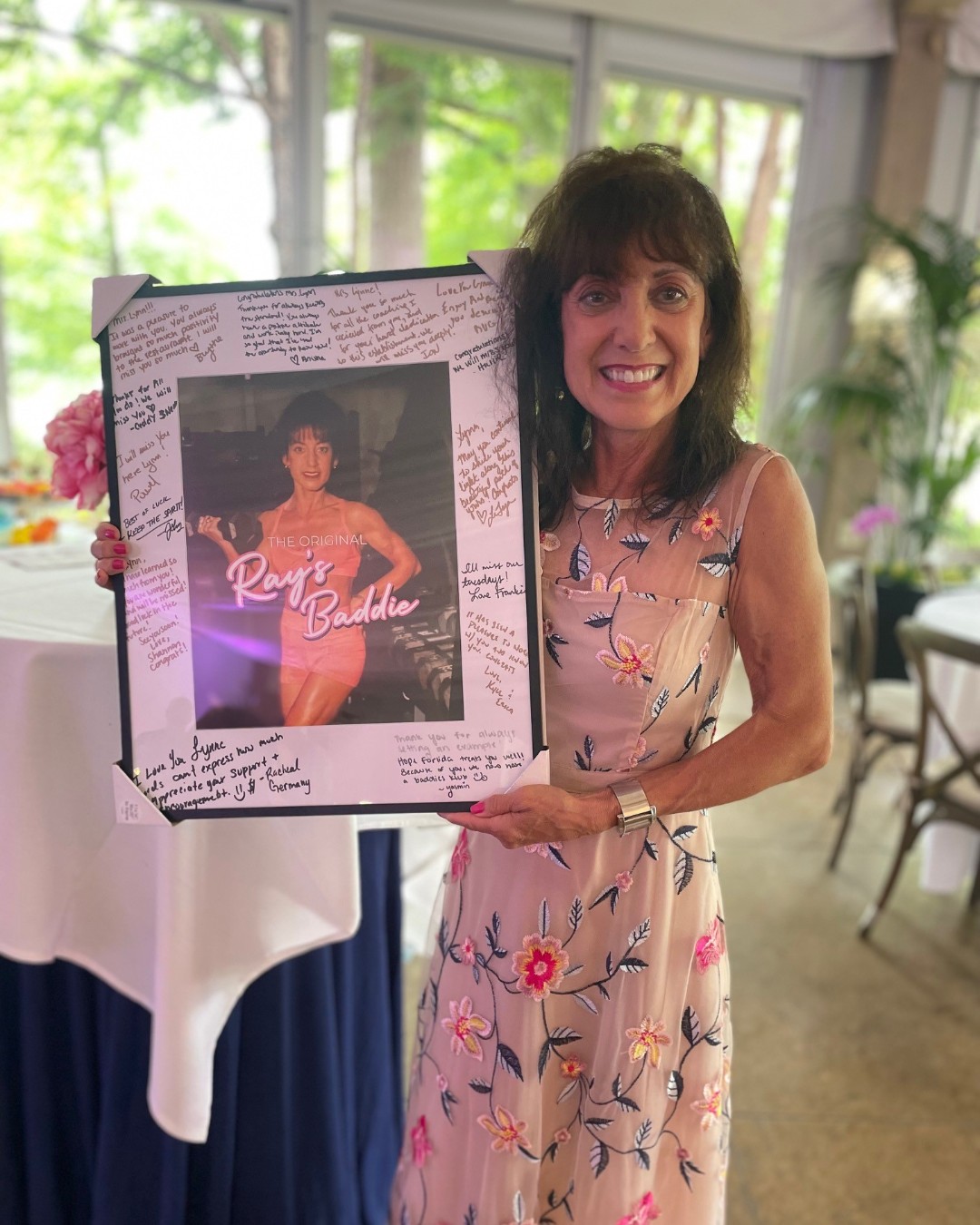Lynne Gomez smiles as she holds a framed photo of herself when she was younger and lifting weights. Text on the photo reads: The original Ray's Baddie. Around the photo is white space where many people have left nice messages for Lynne.