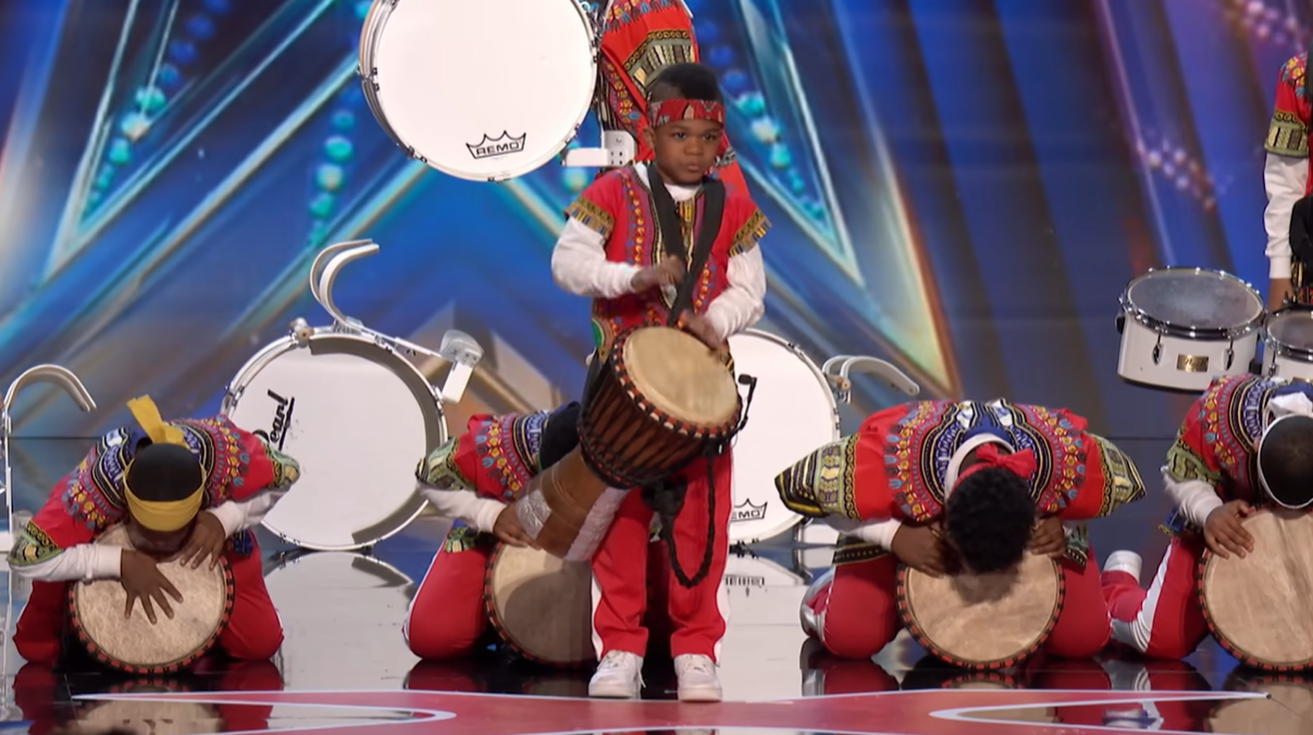 Chioma Narcisse-Williams starts to drum as his fellow drummers kneel and stand behind him with their own drums. They are on the AGT stage.