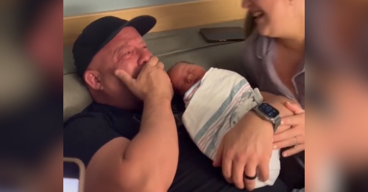 Close up of a man sitting as he holds a newborn. While one arm holds the baby, with the man’s other hand, he covers his mouth, his face red and his eyes full of happy tears. His daughter can be partially seen as she sits next to him and leans closer, smiling wide.