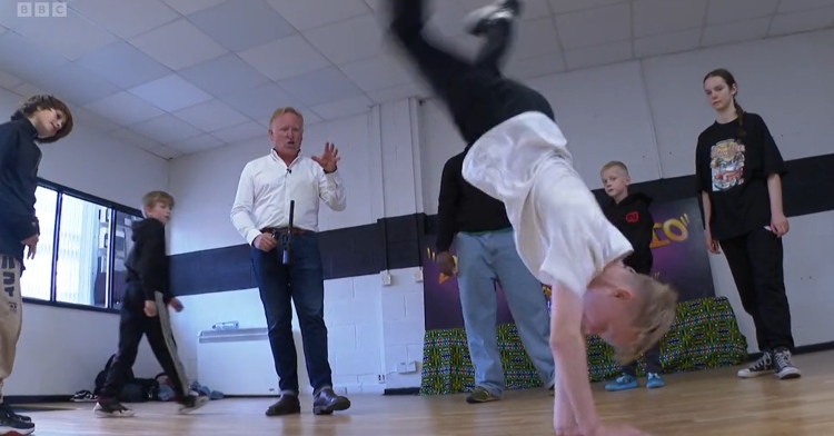 A young boy is mid-handstand while breakdancing. Kids his age, as well as a shocked BBC reporter, stand around and watch.