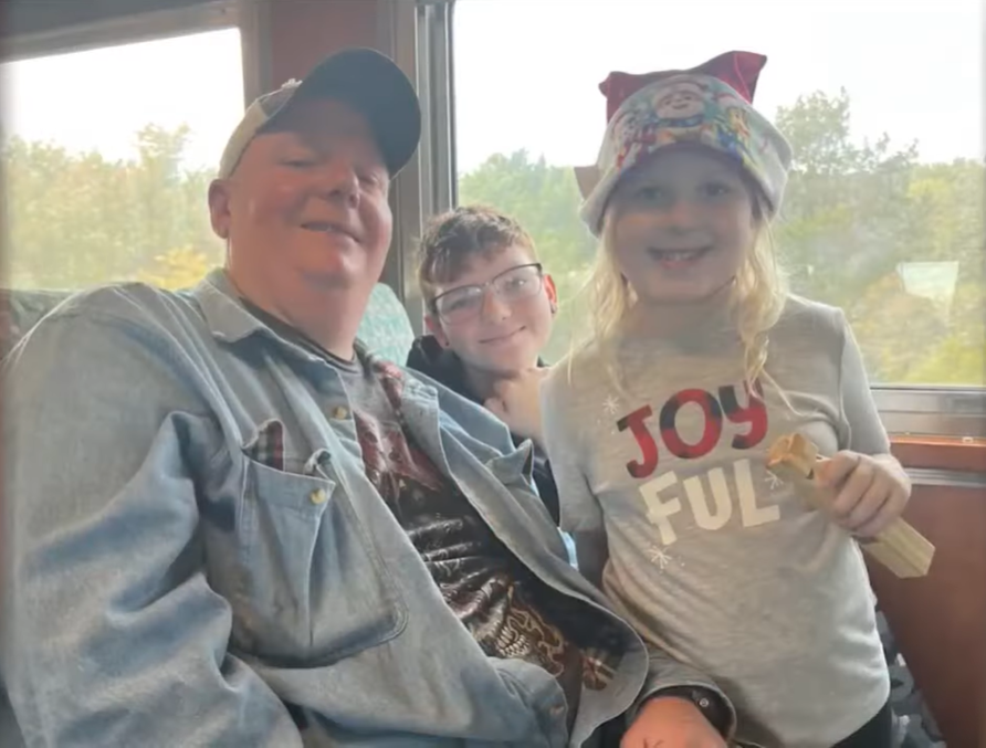 Chris Meffen smiles and poses with two young children on a train.