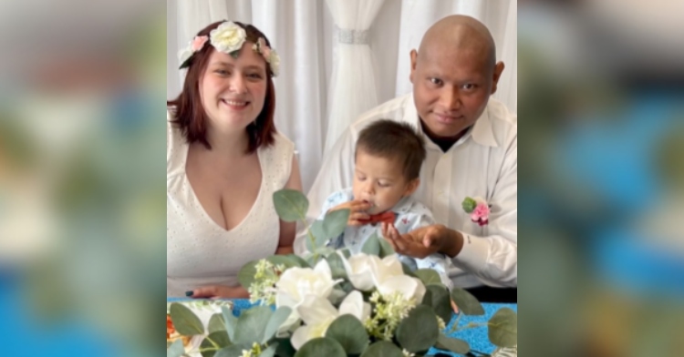 Bride and groom smile at a table with their 2-year-old son at their wedding reception at a hospital.
