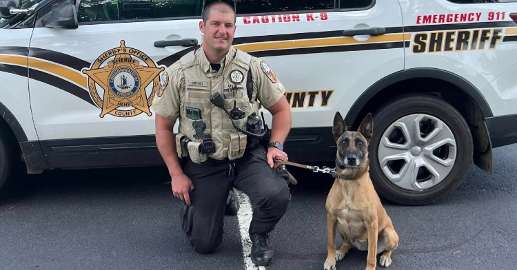 Sgt. Corey Waddell kneels on one knee, posing for a photo with K9 Raja as he holds her leash. They're posing in front of the sheriff's car.