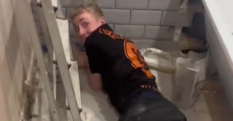 A man lays on his stomach in a bathroom, working. He looks back, eyes wide, and is clearly startled.