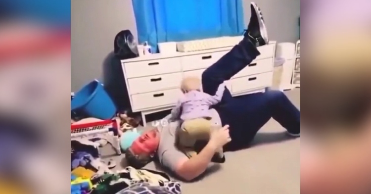 A dad lays on his back on the floor with one leg in the air. His toddler stands over him “attacking” as he grabs the leg that’s in the air.