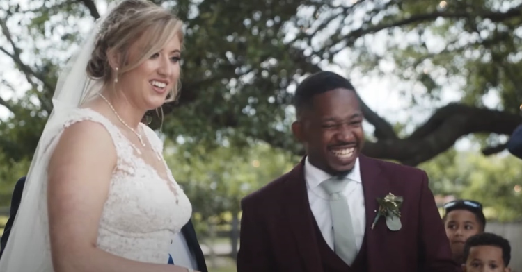 Bride smiles as the groom laughs, both of them looking at something not seen in the image.