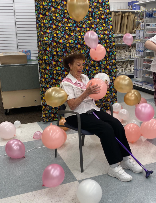 Jayne Burns gives a surprised smile as she sits in a chair in the fabric department of Joann. She's wearing a birthday sash for her 100th birthday and balloons in white, peach, pink, and gold cascade all around her. She's catching a peach one with her hands. A walking cane rests between her legs.