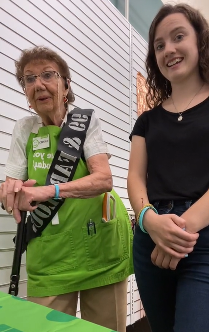 Jayne Burns smiles as she stands next to co-worker Maggie HusVar. Jayne is wearing glasses and a birthday sash to celebrate 99 years.