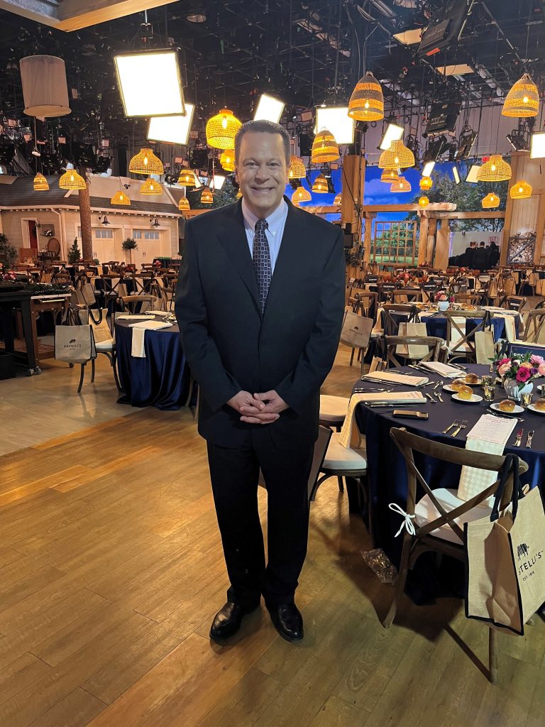 David Venable smiles wide while on a QVC set after having lost weight. Beautifully decorated tables are all around him. He's wearing a suit and tie.