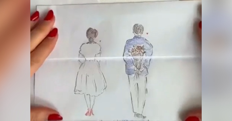 A drawing of a man and woman on a piece of paper. The view is of them from behind. The woman is wearing a white dress that goes past her knees and red shoes. The man is wearing what appears to be either a blue shirt or jacket. He’s holding a bouquet of red flowers behind his back. The artists hands can be partially seen holding down the paper.