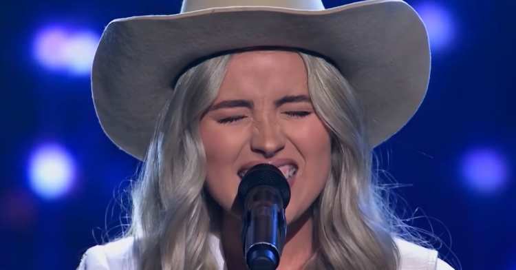 Close up of a blonde woman in a cowboy hat singing into a mic with passion. Her eyes are closed and her noise is scrunched up.