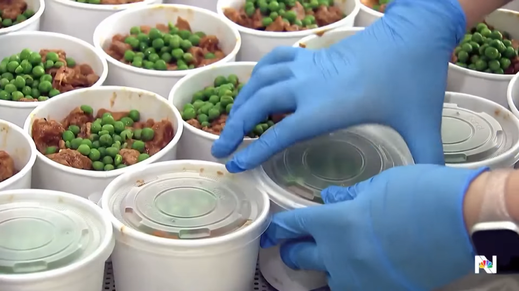 Close up of a person wearing gloves putting lids on food. Inside the containers appears to be some type of meat with peas on top.