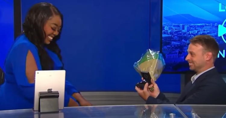 Cornelia Nicholson, a weekend reporter, smiles and looks down shyly as she sits at her desk. Across from her on one knee is her boyfriend and reporter, Riley Nagel. He's smiling as he holds out a bouquet of flowers and a small box containing an engagement ring.