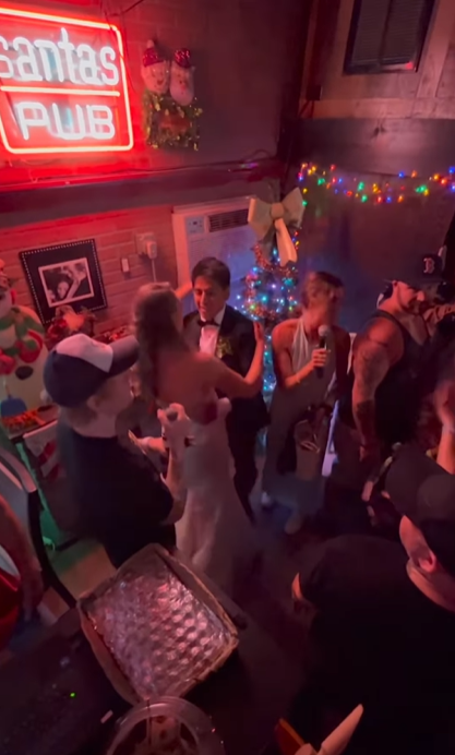 Ed Sheeran chante dans un micro, faisant la sérénade d'un homme et d'une femme vêtus d'une robe de mariée et d'un costume.  Ils sont au Santa's Pun à Nashville.