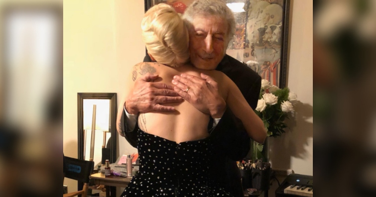 View of Lady Gaga and Tony Bennett hugging. The back of Gaga can be seen as Bennett smiles and hugs her tightly.