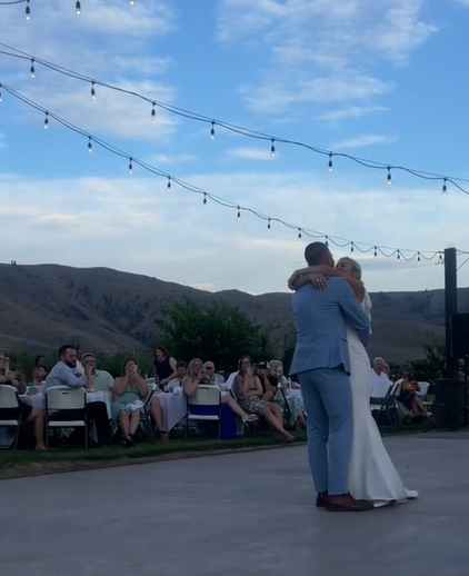 Ivy and her husband slow dance after their wedding. They're dancing outside on a designated dance floor. Lights hang above them and people sitting at chairs sit nearby, watching.