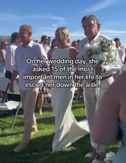 View from the front of Ivy being walked down the aisle at her outdoor wedding. Two men stand at each side with their arms intertwined with Ivy's. Text on the screen reads "On her wedding day, she asked 15 of the most important men in her life to escort her down the aisle."