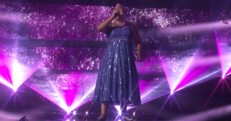 Lavender Darcangelo wears a sparkly blue dress while singing for the second time on the "America's Got Talent" stage. The stage itself is glowing with various purple lights.