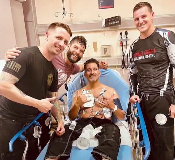 Plymouth police Stgt. Donny Reddington, Det. David Ross, and  firefighter Nick Robbins smile as they stand next to George who is sitting in a hospital bed. George smiles and gives two thumbs up despite being hooked up to several machines.