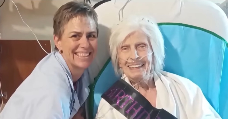 Seana and Marilyn smile and pose for a photo. Marilyn is in a hospital bed with tubes in her nose. She is wearing a birthday sash.