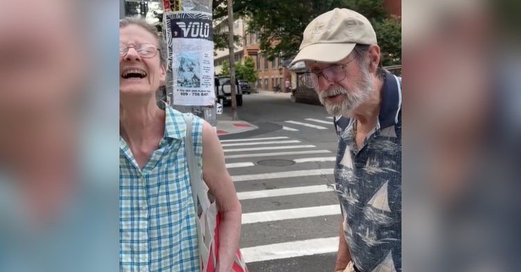 A woman throws her head back with laughter and as her eyes closed. A man, her husband, stands next to her and talks.