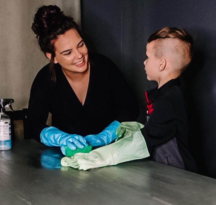 Brogan Ingram smiles with her son at a table. They're both wearing gloves as they clean.