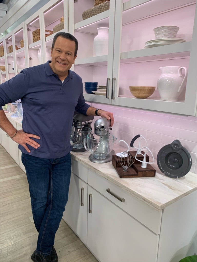 David Venable after losing weight. He looks healthy and happy as he smiles in a QVC kitchen. One hand is on a mixer and the other is on his hip.