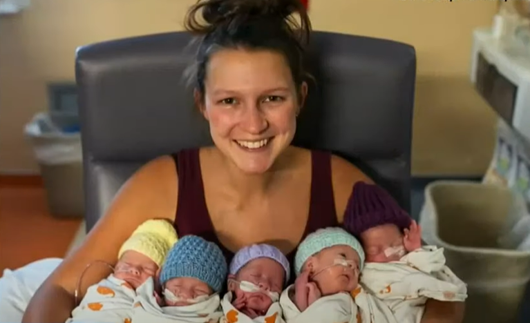 Stephanie Freels smiles wide at the camera as she holds her five newborns in her arms. We can see each baby's face and all but one has their eyes closed. They're each wearing a different colored hat which all appear to be a knitted.