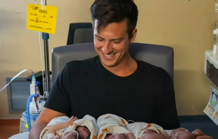 Graham Freels smiles wide as he sits in a chair, looking at his five newborns in his lap and arms.