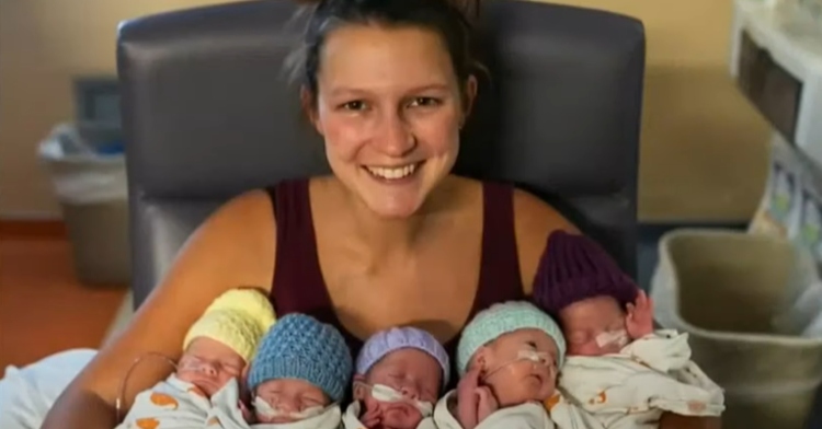 Stephanie Freels smiles wide at the camera as she holds her five newborns in her arms. We can see each baby's face and all but one has their eyes closed. They're each wearing a different colored hat which all appear to be a knitted.