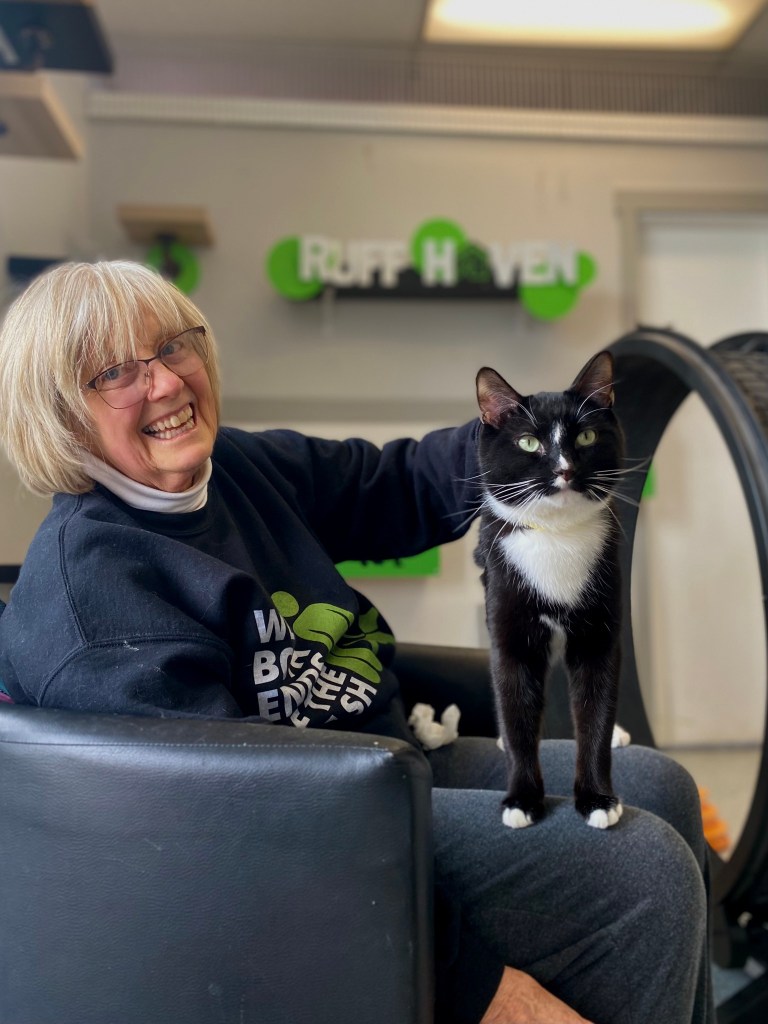 A woman smiles as she sits in a chair. She is wearing a Ruff Haven sweatshirt. A black and white cat stands on her lap.