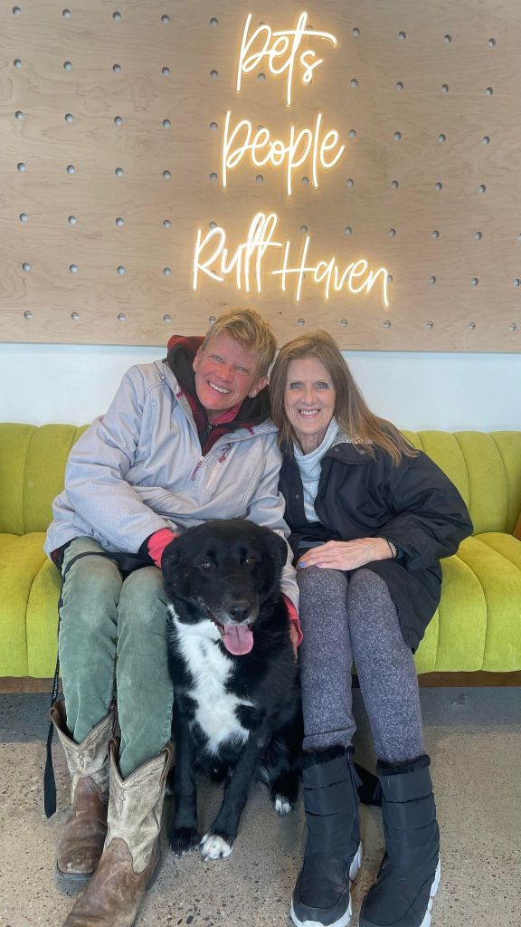 Two people smile as they sit on a green couch inside Ruff Haven. A large black and white dog happily sits between them on the floor, tongue out. On the wall behind them are neon letters that read "pets. people. ruff haven."