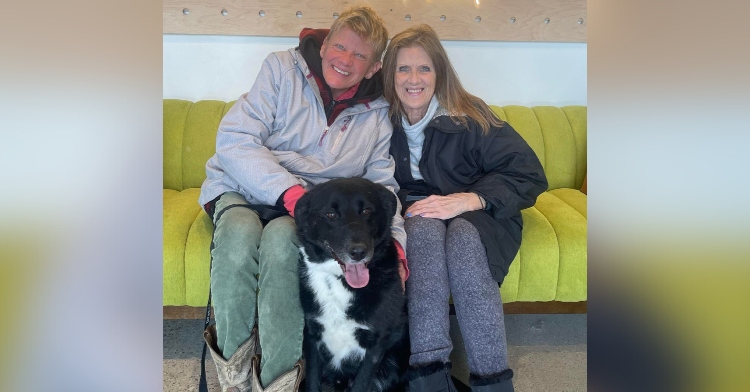 Two people smile as they sit on a green couch inside Ruff Haven. A large black and white dog happily sits between them on the floor, tongue out.