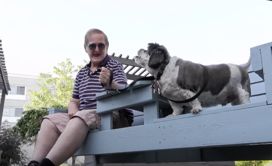 Garry Remick and his 13-year-old dog, Taz, sit outside on a wooden couch. Garry is holding Taz's leash as he smiles and looks over at him. Taz holds his head up and barks.