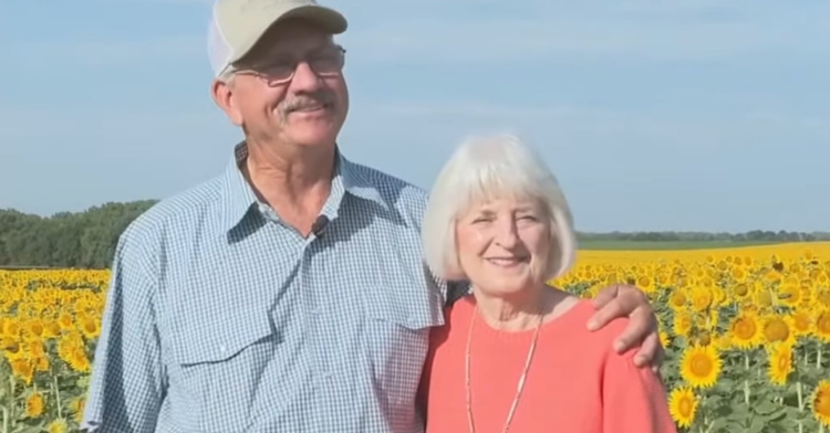 Lee and Renee smile as they stand in their field of sunflowers. Lee has an arm around Renee's shoulder.