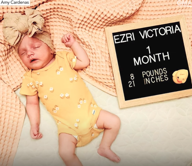 Top-down view of baby Ezri sleeping. She's dressed adorably in a yellow outfit with a cute wrap on her head. A sign next to her reads "Ezri Victoria. 1 month. 8 pounds. 21 inches."