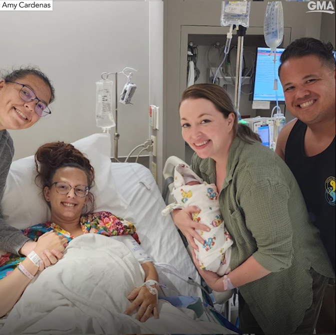 Kelsey and Neva Benton and Amy and John Cardenas in the hospital. Kelsey has just given birth to baby Ora and is sitting up in a hospital bed, holding Neva's hand. John and Amy stand on the other side of the bed. Amy is holding baby Ora. All four of them are smiling.