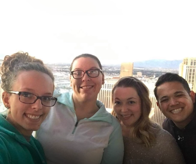 Kelsey and Neva Benton and Amy and John Cardenas smile as they pose for a photo together. Behind them is a view of a city. It appears they are taking the photo on a balcony.