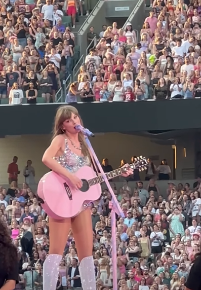 Taylor Swift playing a pink guitar as she sings at the Eras Tour.