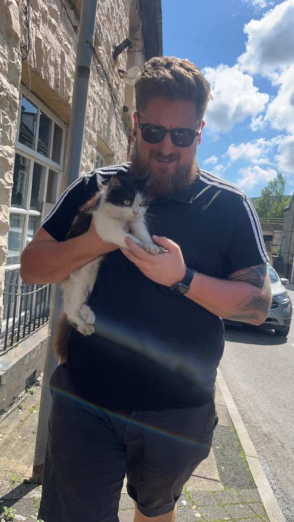 Man named Tom Hutchings smiles as he walks while holding a black and white cat named Gizmo in one hand. With the other hand he's holding her paws. They're outside.