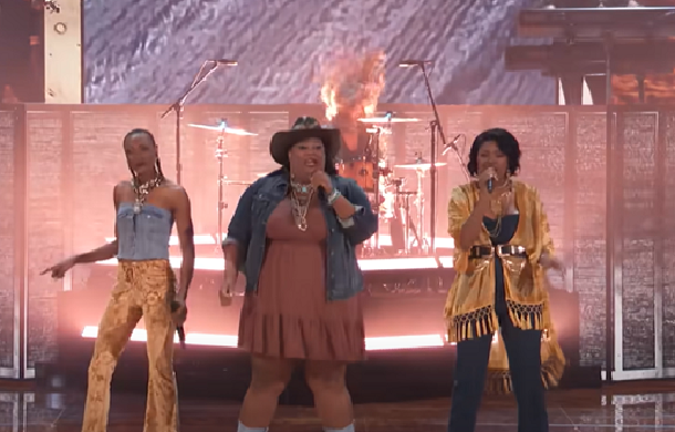 Chapel Hart Band on the AGT stage. Image shows the three female bandmembers singing with a stage-effect flame behind them.