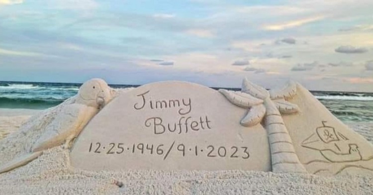 Image shows a sand creation featuring a tribute to the late Jimmy Buffet with his dates of birth and death. A pirate ship is carved into the sand on the right and a parrot on the left. Words of wisdom.