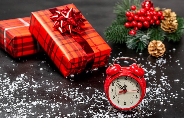 Image shows two packages wrapped in red, plaid holiday papers, a sprig of holly berries, some pine garland, and pinecones with a wind-up clock displaying 11:53.