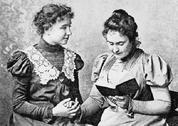 Image shows Helen Keller, seated, with the hand of her lifetime companion in her own as they converse using sign language. Annie Sullivan, reads from a book.