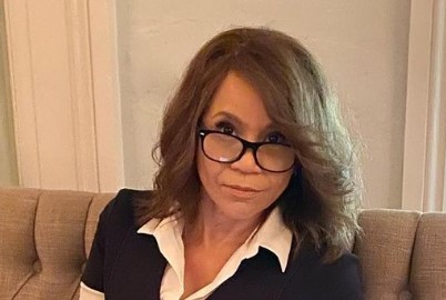Rosie Perez continues inspiring young and old. This image shows her seated on a sofa, wearing glasses that are pushed down on her nose slightly as she peers over the frames.
