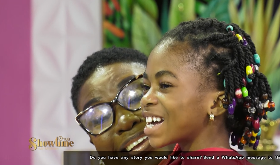 Abigail Adjiri smiles wide, hearing aid in her ears, as she counts to 10 with the "Onua Showtime with Nana Ama McBrown" audience. Abigail is sitting on someone's lap.