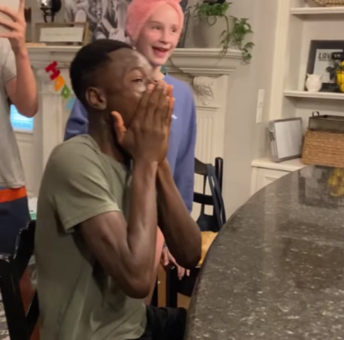 A young man excitedly covers his face with his hands as he sits at a kitchen counter. A younger person stands nearby, mouth open from excitement.