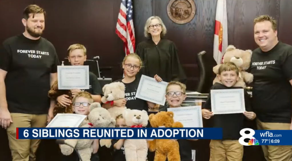 Two men smile along with a judge and their newly adopted children. There are six of them and they are all biologically siblings. The kids hold up their adoption papers and teddy bears.