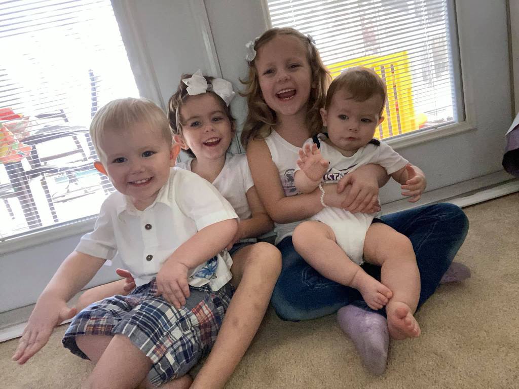 Four young siblings smile as they sit on the floor and on each others' laps.